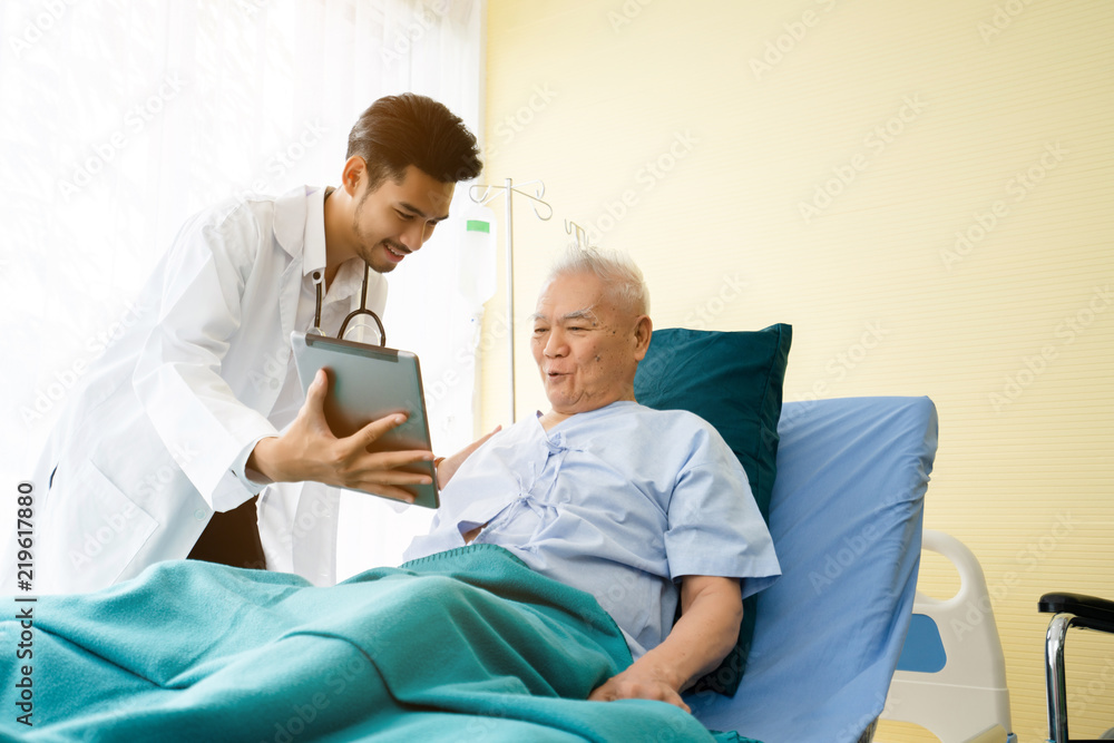 Doctor is standing by to explain the treatment information via the tablet to an elderly patient lying in bed in a private sickroom. The treatment program is suitable for people to socialize elderly. Doctor is standing by to explain the treatment information via the tablet to an elderly patient lying in bed in a private sickroom. The treatment program is suitable for people to socialize elderly.