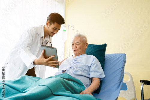 Doctor is standing by to explain the treatment information via the tablet to an elderly patient lying in bed in a private sickroom. The treatment program is suitable for people to socialize elderly.