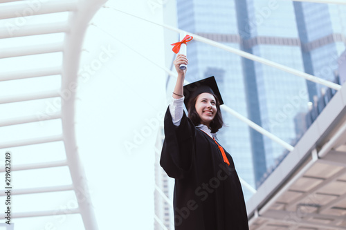 Young woman is happy on her university graduation day.She smiles and holds a graduation certificate in her right hand up in the city.