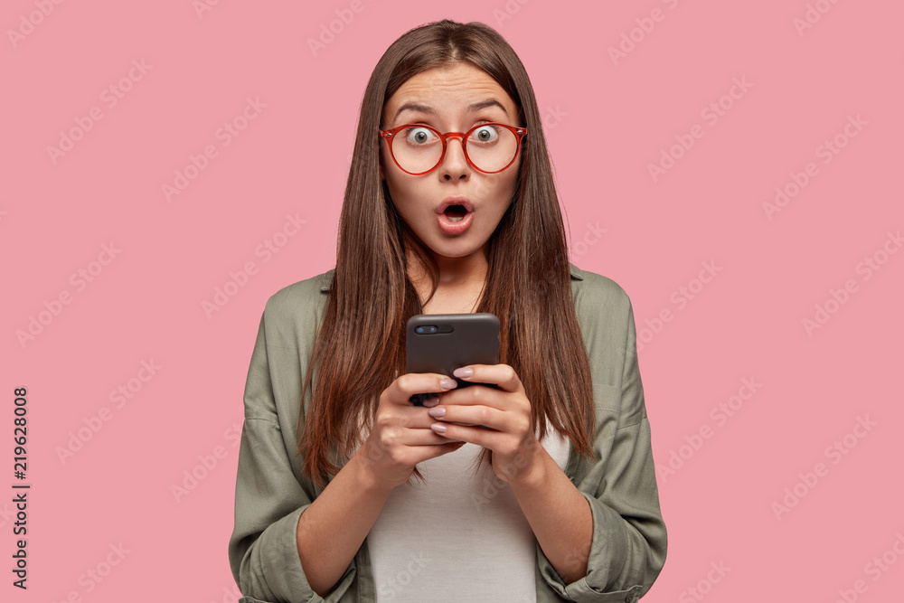 Shocked stupefied attractive girlfriend states with bugged eyes, keeps jaw dropped, forgets about something important while surfes social networks via cell phone, connected to high speed internet Shocked stupefied attractive girlfriend states with bugged eyes, keeps jaw dropped, forgets about something important while surfes social networks via cell phone, connected to high speed internet