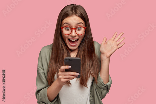 Overjoyed happy excited female glad to recieve text message informing about salary, rejoices good news, stares at mobile phone, gestures actively from happiness, stands indoor alone. Reaction concept