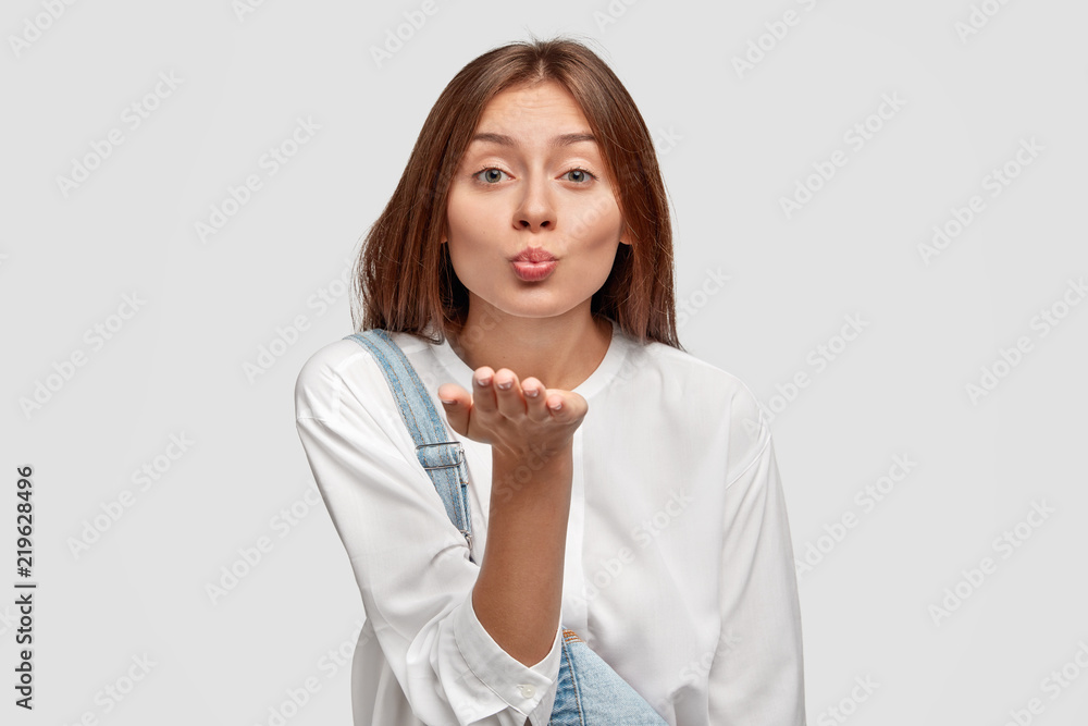 Close up portrait of attractive affectionate girl blows air kiss at camera, stretches hand, has appealing look, dressed casually, isolated over white background, flirts with someone. Body language