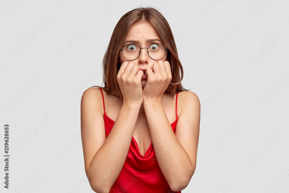 Concerned worried young female has anxious look, bites finger nails, watches horror film before sleep, dressed in red nightdress, worries beacause of tagic events, isolated over white background. Concerned worried young female has anxious look, bites finger nails, watches horror film before sleep, dressed in red nightdress, worries beacause of tagic events, isolated over white background.