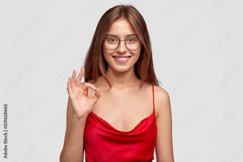 Female adult shows okay gesture, demonstrates that everything is good, has natural smile, dressed in casual domestic outfit, poses against white studio wall. People and body language concept Female adult shows okay gesture, demonstrates that everything is good, has natural smile, dressed in casual domestic outfit, poses against white studio wall. People and body language concept