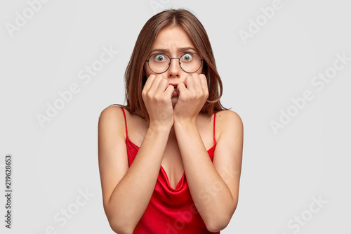 Concerned worried young female has anxious look, bites finger nails, watches horror film before sleep, dressed in red nightdress, worries beacause of tagic events, isolated over white background.