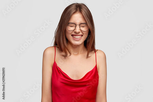 Overjoyed gratified girlfriend laughs happily from funny joke, dressed in elegant red nightdress, squints face from laughter, poses against white background. Optimistic teenager giggles positively