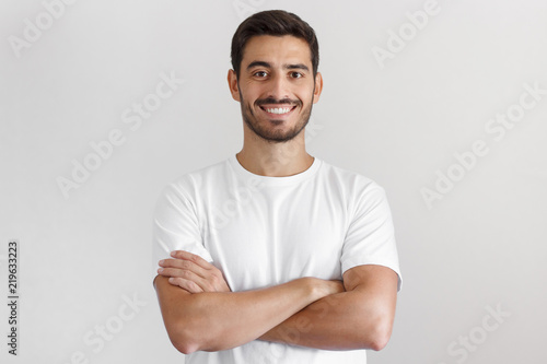 Portrait of smiling handsome man in white t-shirt, standing with crossed arms isolated on gray background photo