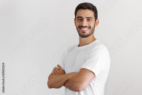 Indoor daylight portrait of young european caucasian man isolated on gray background, standing in white t-shirt with arms crossed, smiling and looking straight at camera photo