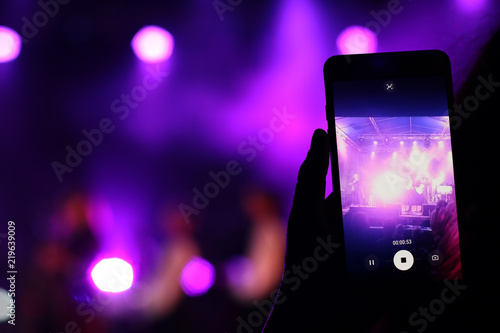 Shooting festival concert on smartphone with place for text. Blurred music stage bokeh background for design. Fans takes picture of scene on phone