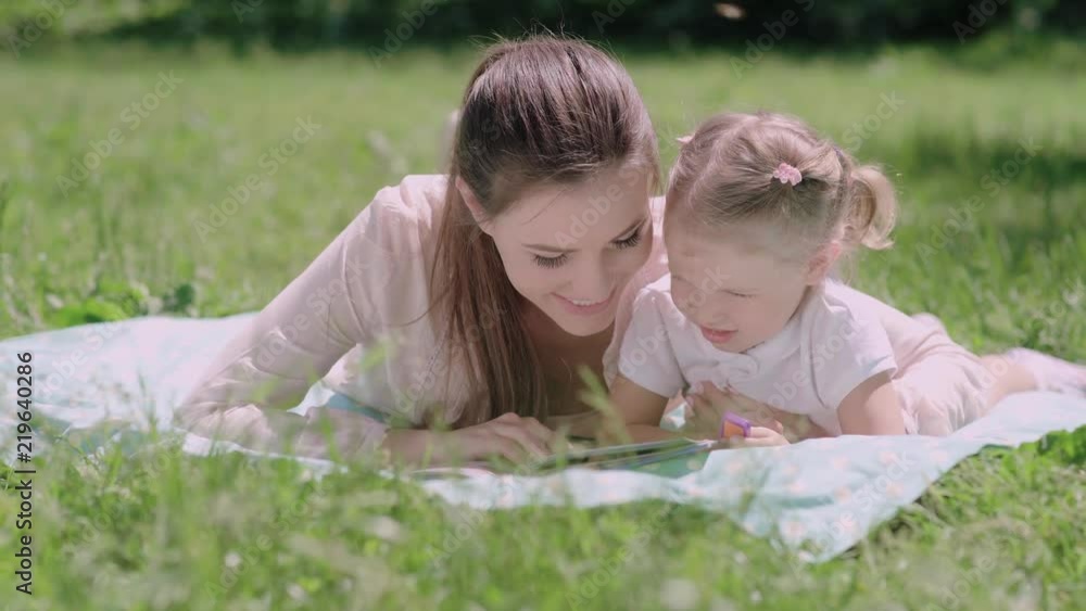 Young Mother And Daughter Reading Book Lying On Grass At Park Young Mother And Daughter Reading Book Lying On Grass At Park