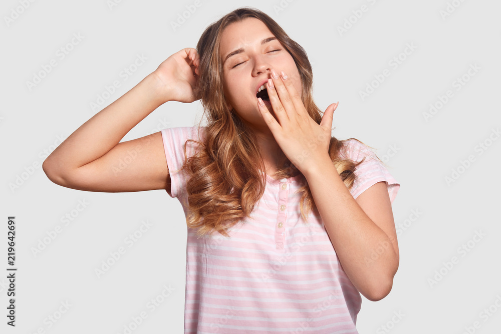 Good morning and awakening concept. Beautiful young woman yawns, covers mouth with hand, stretches awakes early in morning, dressed in casual domestic clothes, isolated over white background. Good morning and awakening concept. Beautiful young woman yawns, covers mouth with hand, stretches awakes early in morning, dressed in casual domestic clothes, isolated over white background.