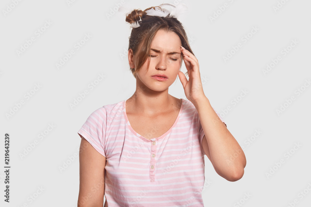 Fatigue woman cant get asleep, has terrible headache, keeps hand on temple, dressed in casual pyjamas, closes eyes from pain, isolated over white background. Weariness and tiredness concept. Fatigue woman cant get asleep, has terrible headache, keeps hand on temple, dressed in casual pyjamas, closes eyes from pain, isolated over white background. Weariness and tiredness concept.