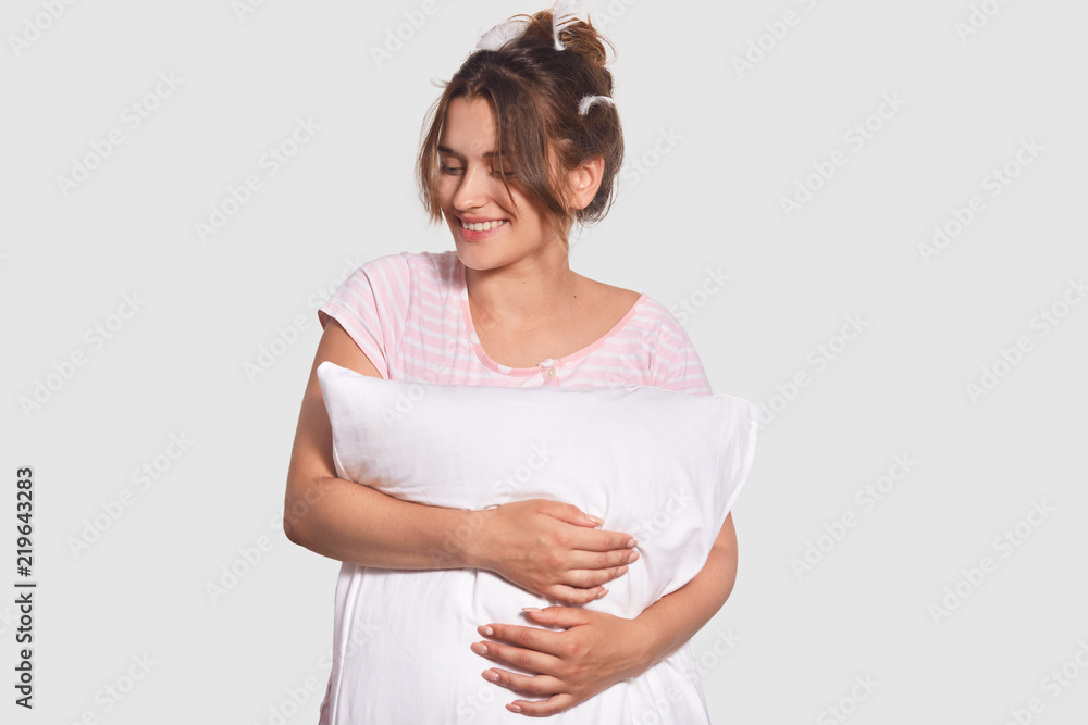 Delighted young female looks positively aside, holds pillow, dressed in casual outfit, has dark combed hair and feathers on head, isolated over white background. People and bed time concept. Delighted young female looks positively aside, holds pillow, dressed in casual outfit, has dark combed hair and feathers on head, isolated over white background. People and bed time concept.