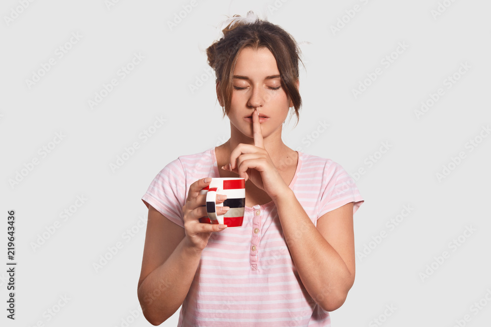 Indoor shot of relaxed young female makes shush gesture, dressed in nighclothes, asks to be quiet early in morning, holds mug with hot coffee or tea, stands against white background. Drinking concept Indoor shot of relaxed young female makes shush gesture, dressed in nighclothes, asks to be quiet early in morning, holds mug with hot coffee or tea, stands against white background. Drinking concept