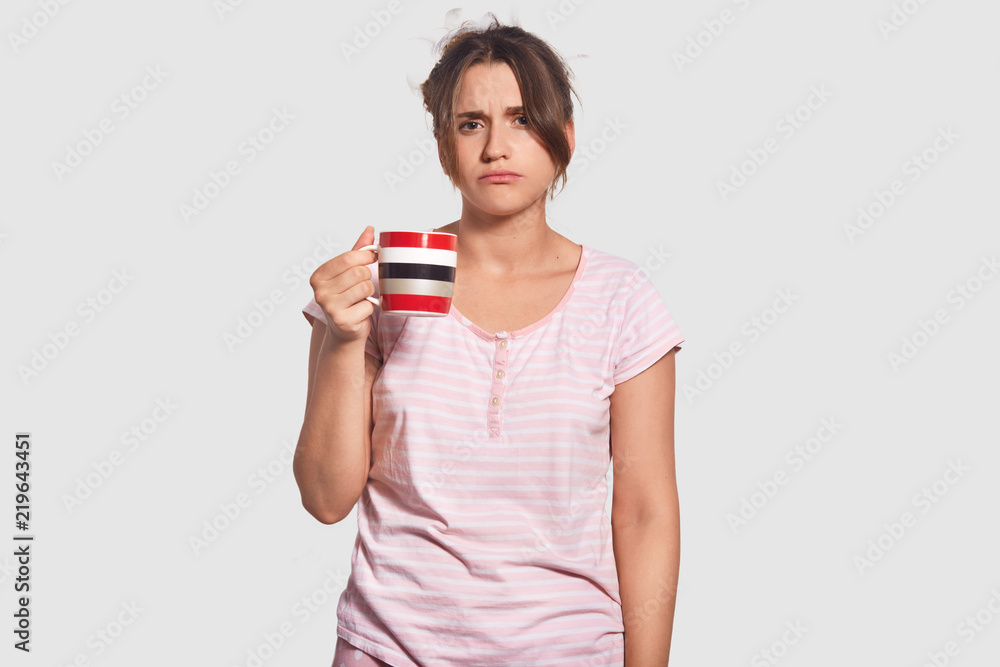 Waist up portrait of unhappy woman has gloomy sleepy expression, tries to wake up, drinks hot coffee, doesnt want to go for work, dressed in domestic clothing, isolated over white background Waist up portrait of unhappy woman has gloomy sleepy expression, tries to wake up, drinks hot coffee, doesnt want to go for work, dressed in domestic clothing, isolated over white background
