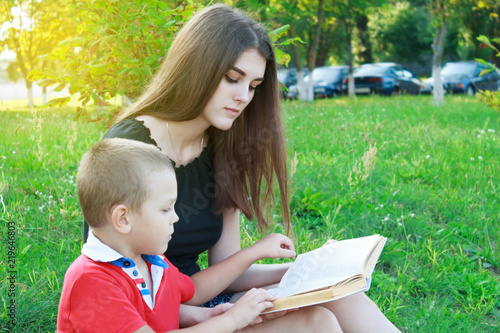 A brother and sister are sitting on the green grass and reading a book.