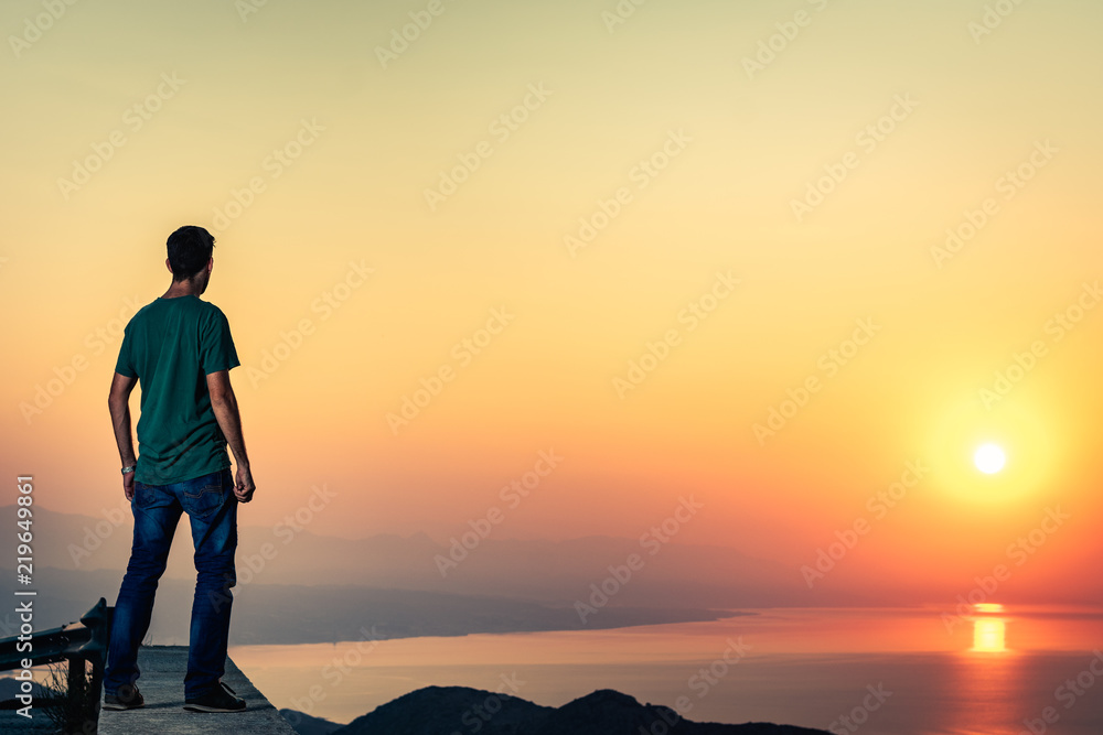 Hombre mirando el amanecer desde arriba de una montaña, sol reflejado en el mar