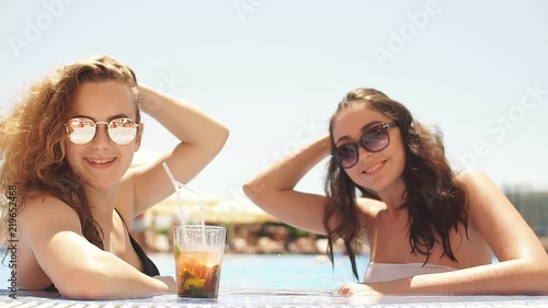 Outdoor portrait of two young cute girlfriends in sunglasses and swimsuits with cocktail glass chilling in swimming pool in tropical resort. Vacation concept. photo