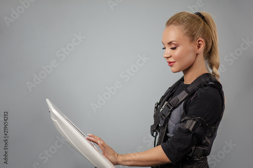 close up side view photo of a beautiful fit fair-haired woman touching the ems device during workout. isolated gray background. sportswoman controlling the training.sport, health, hobby cooncept photo