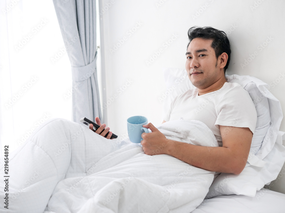Obraz premium Smiling Asian man using smartphone and drinking coffee on bed, man lifestyle on weekend concept.