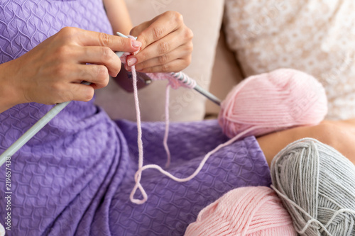 Close-up of woman in a lilac home dress knits a light pink scarf or plaid from natural threads on the bed.
