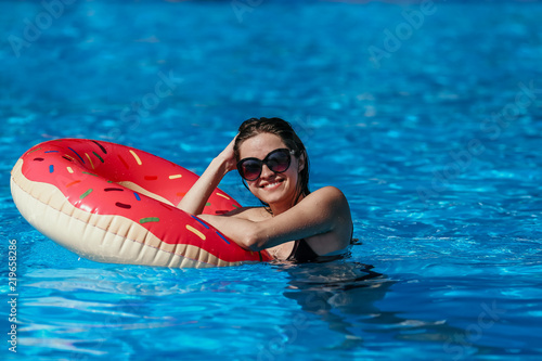 Beautiful young woman with pink donut circle have rest in blue swimming pool 