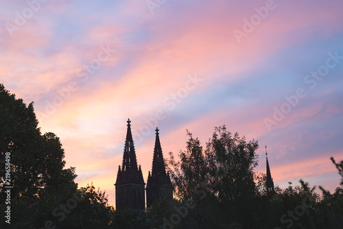 towers Church of St Ludmila Kostel sv. Ludmily in Prague in the distance against the background of the sunset and the pink sky and green forest © YuliiaMazurkevych