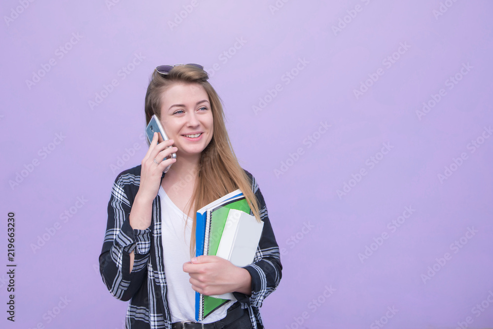 Smiling girl talking on the phone and holding a notebook and books on purple background. Happy student talking on a smartphone and looking away, isolated on a purple background.Copyspace Smiling girl talking on the phone and holding a notebook and books on purple background. Happy student talking on a smartphone and looking away, isolated on a purple background.Copyspace