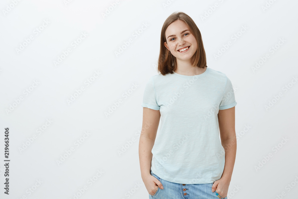 Friendly cute white girl with short haircut and lovely flapped ear smiling joyfully holding hands in pockets and gazing at camera with joyful entertained look posing over white background Friendly cute white girl with short haircut and lovely flapped ear smiling joyfully holding hands in pockets and gazing at camera with joyful entertained look posing over white background