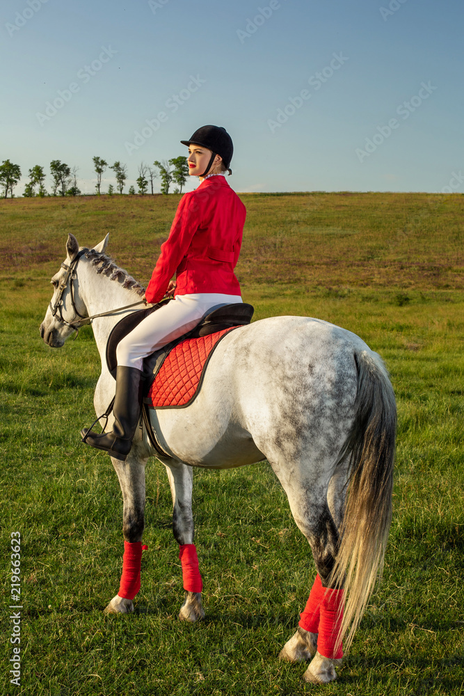 Young woman rider, wearing red redingote and white breeches, with her horse in evening sunset light.