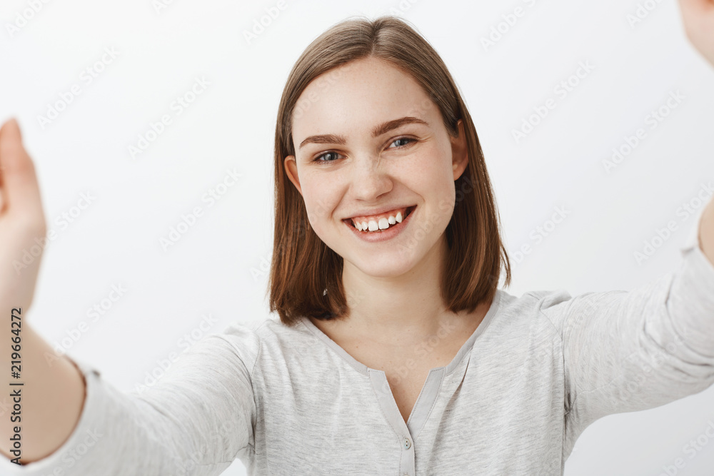 Close-up shot of joyful friendly-looking energized brunette girl in casual outfit pulling hands towards camera as if holding smartphone and taking selfie smiling broadly posing over gray background Close-up shot of joyful friendly-looking energized brunette girl in casual outfit pulling hands towards camera as if holding smartphone and taking selfie smiling broadly posing over gray background