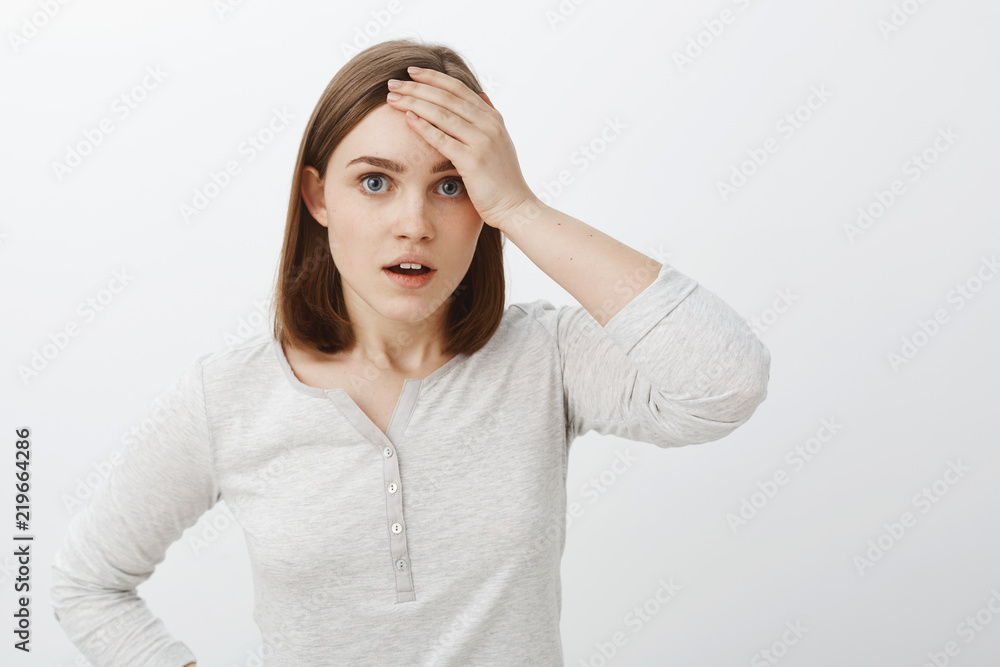 Why me god. Portrait of embarrassed and worried young female student standing bothered and concerned over grey wall in casual white blouse holding palm on forehead complaining on hard life Why me god. Portrait of embarrassed and worried young female student standing bothered and concerned over grey wall in casual white blouse holding palm on forehead complaining on hard life