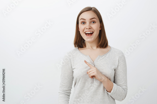 Waist-up shot of enthusiastic energized cute girl reacting to awesome offer of friend going sit in cafe smiling joyfully with amused expression gazing at camera and pointing at upper left corner © Cookie Studio