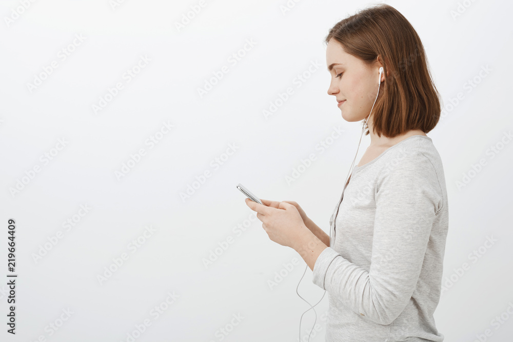 Profile shot of cute sociable young caucasain female brunette wearing earphones standing against grey wall in casual white blouse holding smartphone smiling while texting friend on her way to party