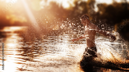 a young man with a bare torso in black shorts running through the water with splashes against the sunset