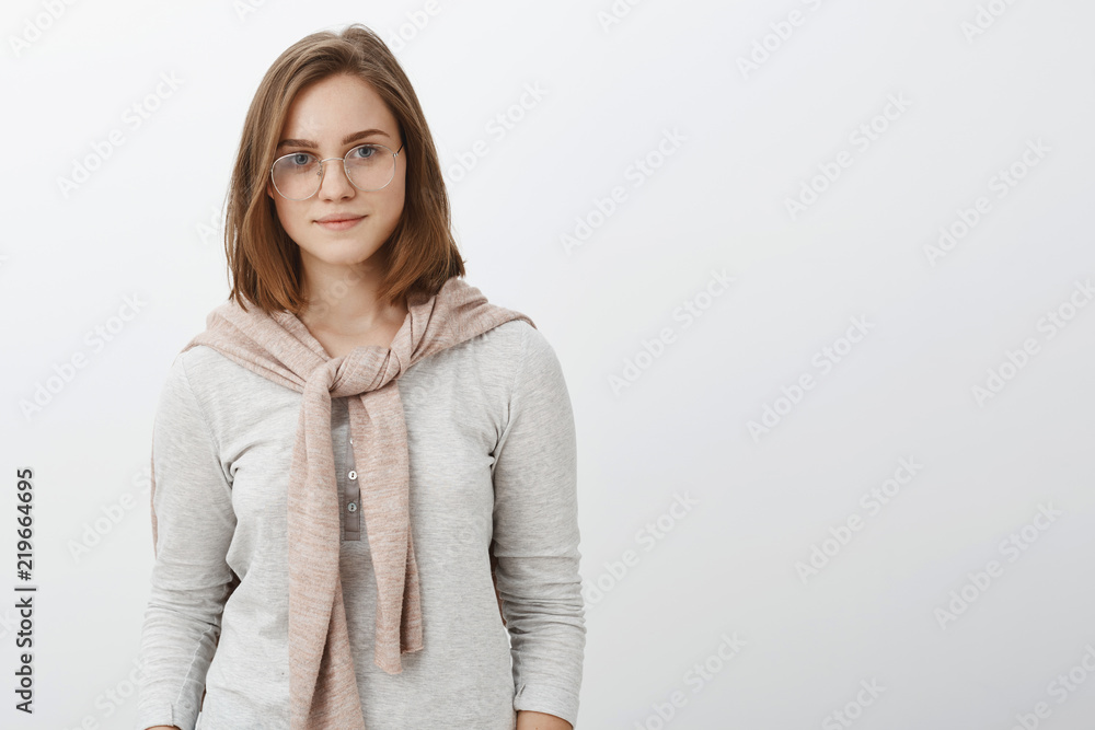 Waist-up shot of attractive stylish hipster girl in glasses and sweatshirt on shoulders smiling self-assured at camera working on new project standing confident and assured over gray background Waist-up shot of attractive stylish hipster girl in glasses and sweatshirt on shoulders smiling self-assured at camera working on new project standing confident and assured over gray background