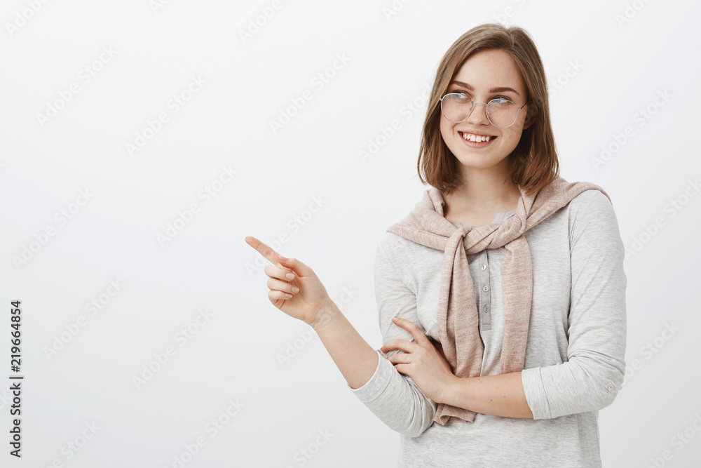 Waist-up shot of creative attractive brunette female coworker in glasses and pullover tied over neck gazing and pointing left with delight and interest promoting cool item over gray background Waist-up shot of creative attractive brunette female coworker in glasses and pullover tied over neck gazing and pointing left with delight and interest promoting cool item over gray background