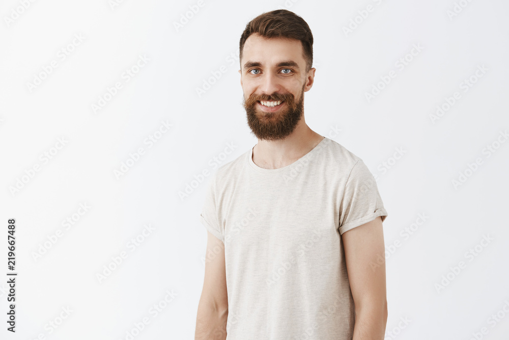 Fototapeta premium Happy creative and young bearded male coworker with stylish hairstyle in white casual t-shirt smiling joyfully standing over gray background talking about new company concepts creating new ideas