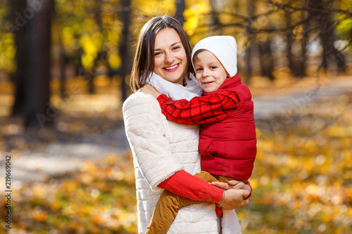 Cute,happy, white boy in red shirt smiling and hugging with his mom among yellow leaves. Little child having fun with mother in autumn park. Concept of friendship between son and parents, happy family