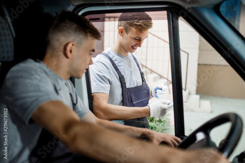 A young handsome guy wearing uniform is looking out of the car window. Another worker wearing uniform is holding the clipboard. House move, mover service.