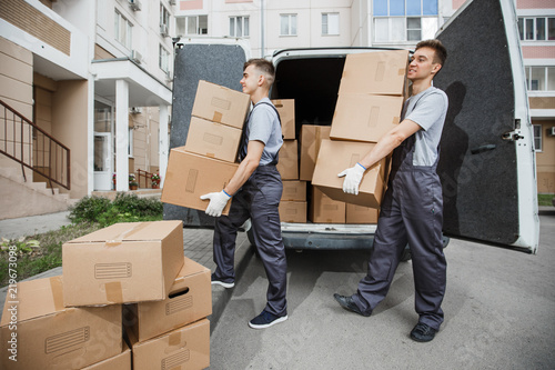 Two young handsome smiling workers wearing uniforms are unloading the van full of boxes. The block of flats is in the background. House move, mover service.