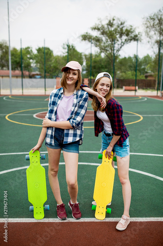 Two pretty smiling blond girls wearing checkered shirts, caps and denim shorts are standing on the sportsfield with bright longboards in their hands.