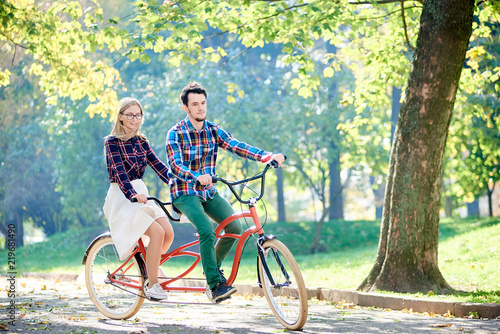 Young active smiling traveler couple, handsome bearded man and attractive blond woman riding together tandem double bike along path in lit by bright sun beautiful park under tall trees.