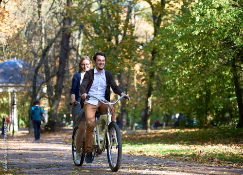 Young active tourist couple, handsome bearded man and attractive blond long-haired woman cycling together tandem double red bike by sunny alley with golden leaves on tall trees background.