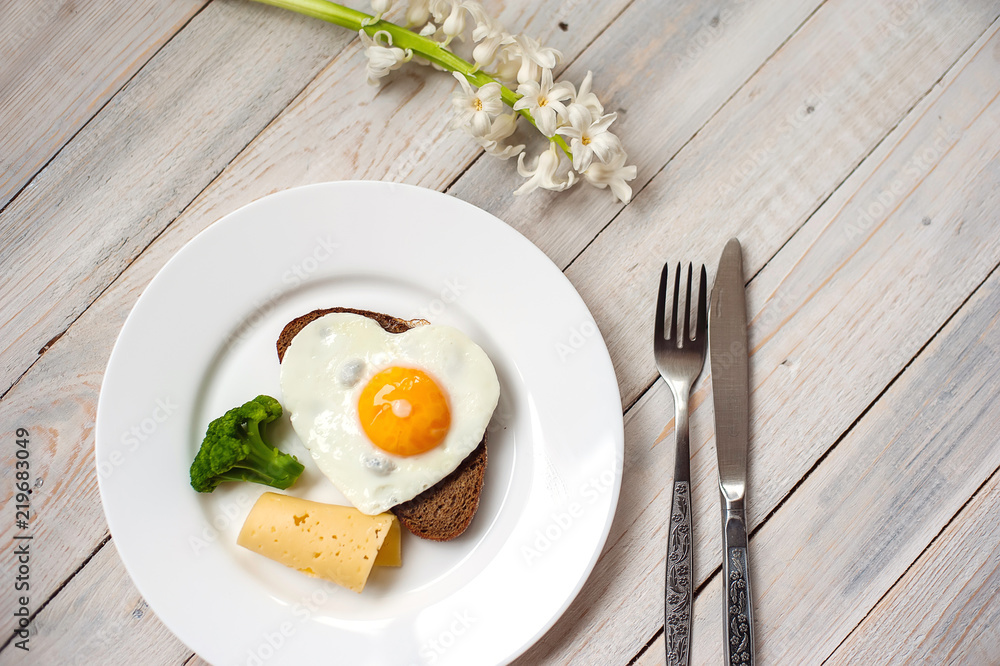 The fried egg in the form of heart with vegetables and bread on a white plate The fried egg in the form of heart with vegetables and bread on a white plate