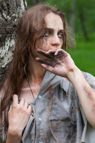 Beautiful young dirty mad and manic looking girl wearing torn clothes and smeared with mud and dried blood stands and huddles to a tree and wipes her face with her hand in the forest.