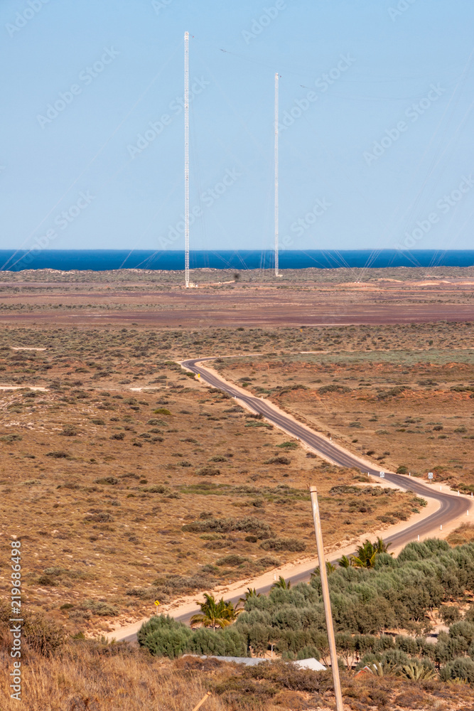 Exmouth, Western Australia - November 27, 2009: Winding road through bush land leading to tall white antennas in the back of nearby Naval base. Horizon is deep blue Indian ocean under light blue sky.