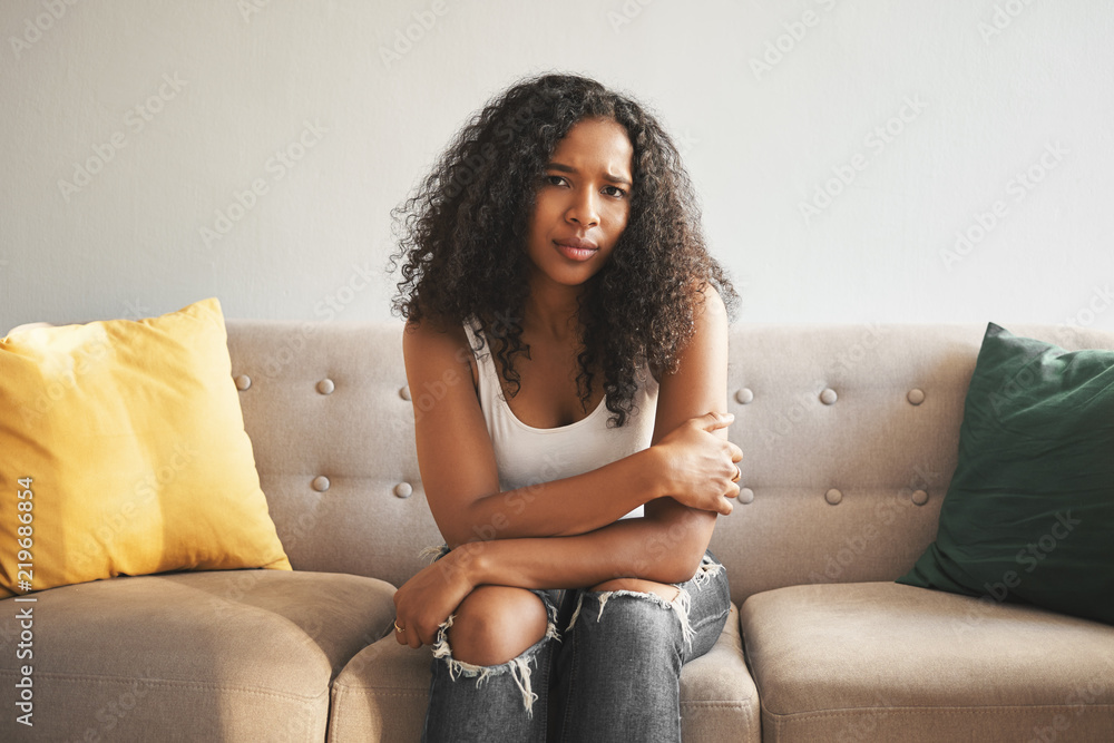 Indoor shot of beautiful fashionable young mixed race woman with Afro hairstyle sitting on couch at home, frowning, having worried sad look, suffering from stomach cramps or feeling lonely and upset Indoor shot of beautiful fashionable young mixed race woman with Afro hairstyle sitting on couch at home, frowning, having worried sad look, suffering from stomach cramps or feeling lonely and upset