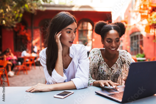 Pretty girl amazedly looking in laptop while working with female friend together in courtyard of cafe