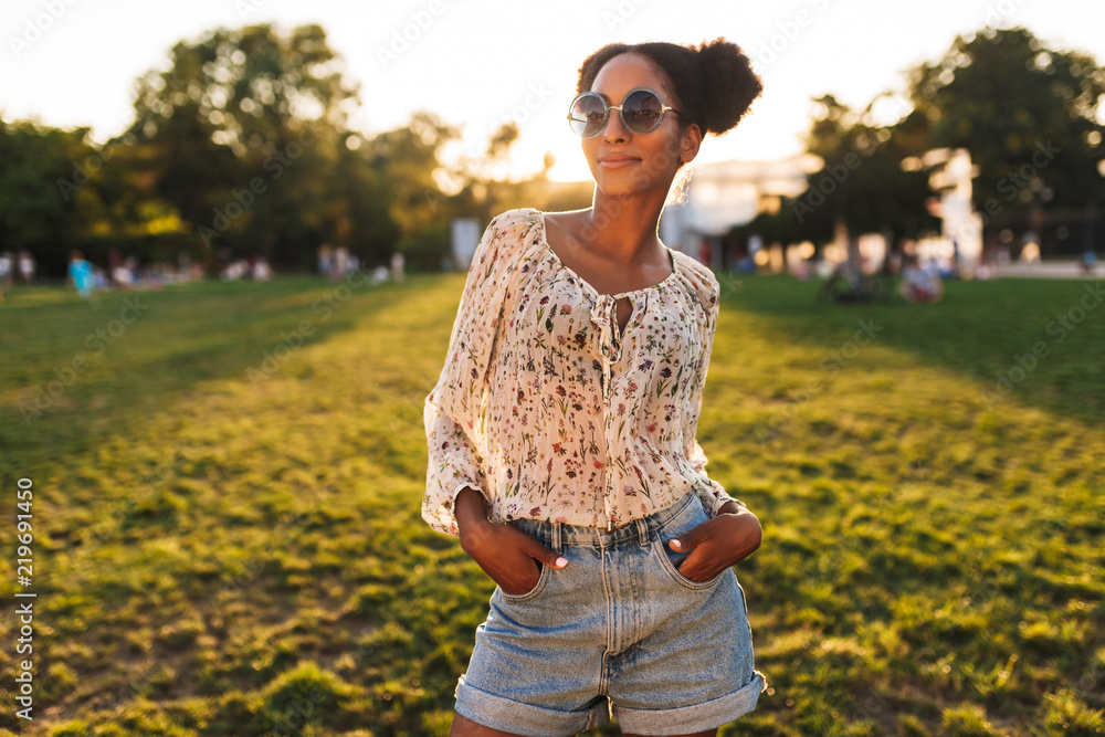 Fototapeta premium Young beautiful african woman in sunglasses dreamily looking in camera while spending time in city park